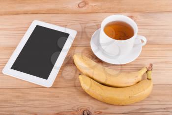 Digital tablet cup of tea and bananas on a wooden table.