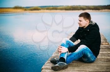 Young handsome man sitting on wooden pier in autumn day, relaxing,  thinking, listening. Casual style - jeans, jacket