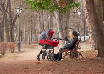 a woman with a baby carriage rests on a park bench and reading a book