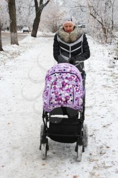 Woman with baby in perambulator walking on the winter road with tree standing in the hoar-frost