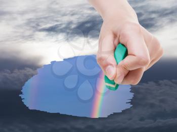 weather concept - hand deletes storm clouds on sky by rubber eraser from image and rainbow are appearing