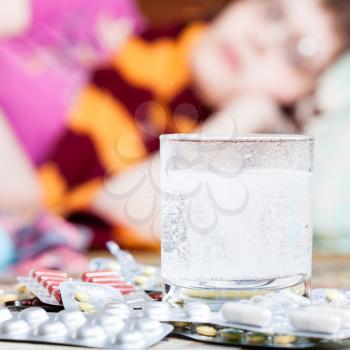 dissolving drug in glass with water and pills on table close up and sick woman with scarf around her neck on sofa in living room on background