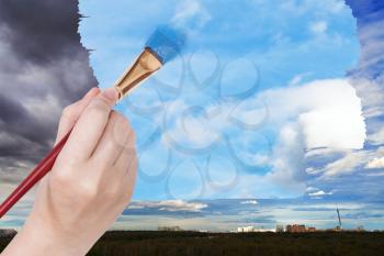 nature concept - seasons and weather changing: hand with paintbrush paints blue sky on rainy clouds over city