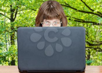 girl with spectacles looks over cover of open laptop with green summer forest on background