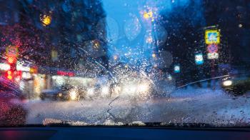 driving in night snowfall in Moscow - unfocused background with view of street in residential quarter in snowy evening through wet windscreen (focus on melting trickle at the bottom of car glass)