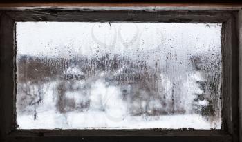 blurry view of russian village through water drops from melting snow on surface of frozen window of rural house in cold winter day (focus on surface of the glass)