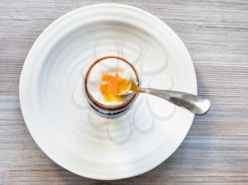 top view of soft-boiled brown egg with spoon in cup on white plate on gray wooden board
