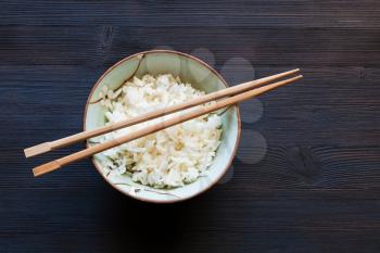 top view of chopsticks above boiled rice in bowl on dark wooden board with copyspace