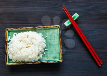 top view of portion of boiled rice on green plate and red chopsticks on rest on dark wooden board