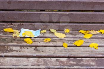 dropped sanitary face mask covered by yellow fallen leaves on shabby wooden bench in city park on autumn day
