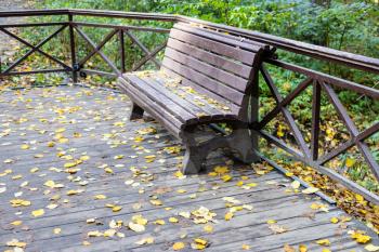 old wooden bench on wooden pavement covered by yellow fallen leaves in city park on autumn day