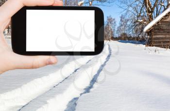 travel concept - tourist photographs of ski track to forest in little village in sunny winter day in Smolensk region of Russia on smartphone with cut out screen with blank place for advertising