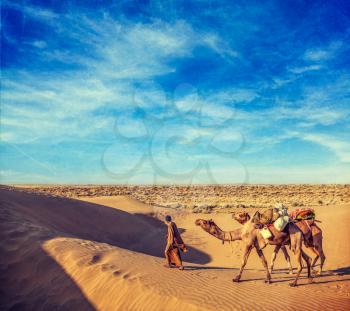 Vintage retro hipster style travel image of Rajasthan travel background - India cameleer (camel driver) with camels in dunes of Thar desert with grunge texture overlaid. Jaisalmer, Rajasthan, India
