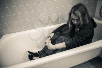 Sad Caucasian teenage girl sitting in empty bath. Monochrome photo. Depression concept