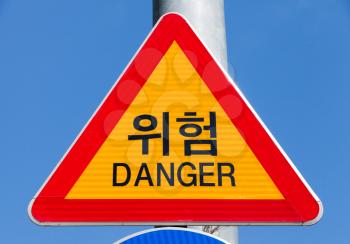 Danger, yellow triangle warning traffic sign with red frame over blue sky with Korean and English text