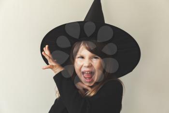 Little grimacing blond European girl in black witch costume over white wall, close-up studio portrait