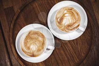 Two cups of cappuccino coffee stand on dark wooden table, close-up top view