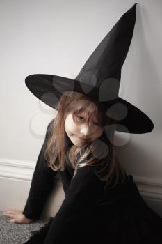 Little blond European girl in black witch costume sitting over white wall, close-up studio portrait