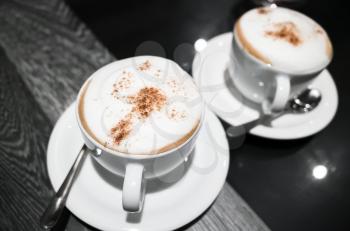 Cappuccino, two cups of coffee with milk foam and cinnamon stand on black table in cafeteria, closeup photo with selective focus