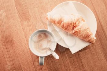 Cappuccino with croissant. Cup of coffee stands on wooden table in cafeteria, closeup photo with selective focus, top view, retro style tonal correction filter effect