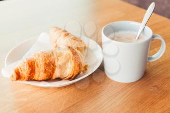 Cappuccino with croissant. Cup of coffee with milk foam stands on wooden table in cafeteria, close up photo with selective focus