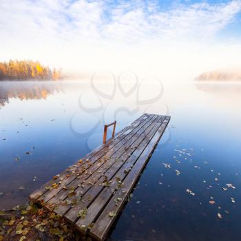 Small wooden pier on still lake in bright autumnal foggy morning