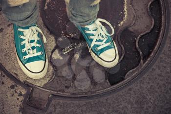 Teenager in sneakers. Feet in gumshoes stand on a rusty urban manhole cover. Closeup photo with selective focus and vintage tonal correction filter, old style 