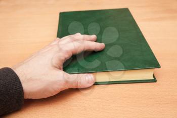 Male hand and book with empty dark green leather cover lay on wooden desk
