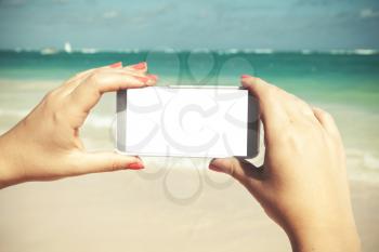 Woman holds smart phone with white empty screen in hands for taking photo on a beach in Dominican republic. Vintage tonal correction photo filter, old style effect