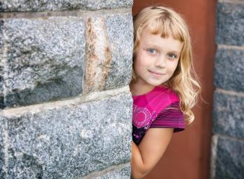 Little blond girl looks out from behind the gray stone wall and smiles