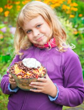 Little blond girl smiles and holds homemade fruit dessert. Outdoor bright portrait