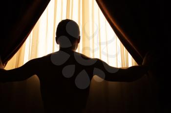 Man in dark room opens curtains on window to the morning sunlight