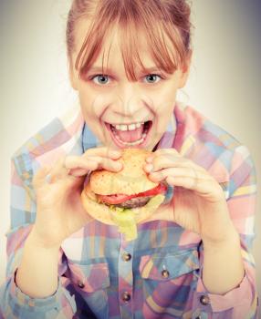 Little blond smiling girl eats hamburger above white wall. Instagram toned effect