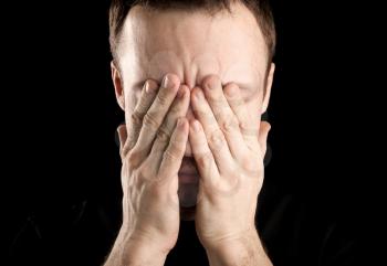 Portrait of young stressed Caucasian man covers his face with hands isolated on black background