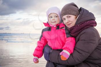 Caucasian family outdoor portrait on winter sea coast, young mother hugs her little baby girl in pink jacket