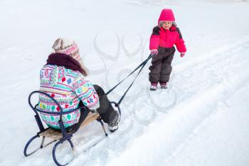 Little baby girl in pink pulling a sled with her big sister on snowy winter road