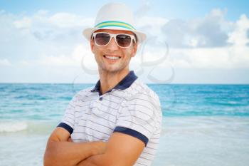Outdoor portrait of young smiling Caucasian man in white hat and sunglasses standing on the Atlantic ocean coast