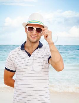 Outdoor portrait of young smiling Caucasian man in white hat and sunglasses standing on the ocean coast