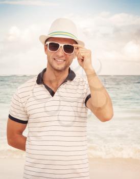 Outdoor portrait of young smiling Caucasian man in white hat and sunglasses standing on the ocean coast. Vintage toned photo with style photo filter effect