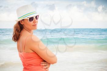 Young happy Caucasian woman in red dress, and sunglasses and white hat. Summer outdoor portrait on the sea coast. Vintage toned photo with warming filter effect