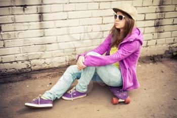 Blond teenage girl in jeans, cap and sunglasses sits on her skateboard near urban brick wall, photo with warm retro tonal correction effect, instagram old style filter
