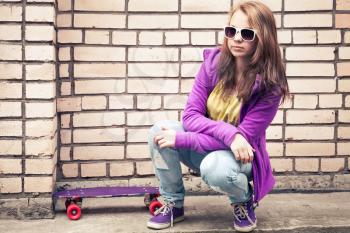 Blond teenage girl in a sunglasses with skateboard sits near urban brick wall, photo with retro tonal correction effect, instagram old style filter