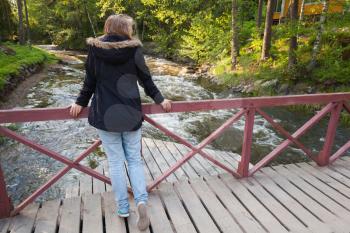 Beautiful blond Caucasian teenage girl stands on wooden bridge across small fast river in Finland, back view