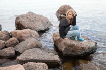 Beautiful blond Caucasian teenage girl sits on coastal stones on the lake coast in Finland