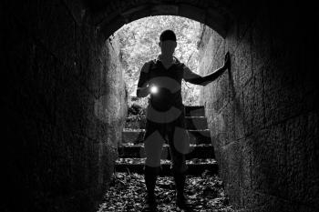Young man with a flashlight enters the stone tunnel and looks in the dark, monochrome photo