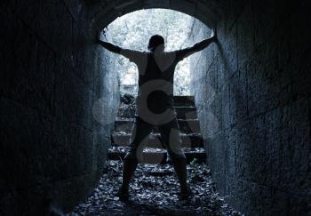 Young man stands in dark stone tunnel with glowing end, blue tonal correction filter