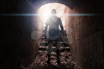 Young man stands in dark stone tunnel with glowing end, warm toned photo with lens glow effect