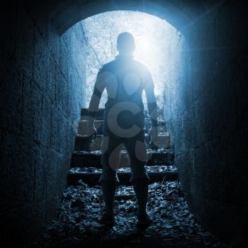 Young man stands in dark stone tunnel with glowing end, blue toned square photo