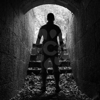 Young man stands in dark stone tunnel with glowing end, black and white square photo
