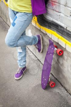 Teenager in blue jeans and gumshoes stands with skateboard near gray brick wall